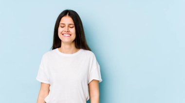 Mavi arka planda izole edilmiş genç beyaz kadın gülüyor ve gözlerini kapıyor, rahatlamış ve mutlu hissediyor..