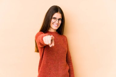 Beyaz tenli genç bir kadın, bej arka planda izole edilmiş, gülümseyen, ön tarafı işaret eden..