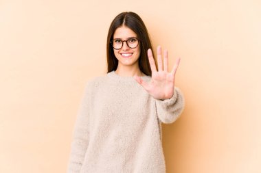 Beyaz tenli genç bir kadın bej arka planda izole edilmiş. Gülümsüyor. Beş numarayı parmaklarıyla gösteriyor..