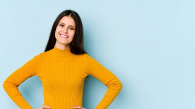 Kafkasyalı genç bir kadın mavi arka planda izole edilmiş. Kendine güvenen, ellerini kalçasında tutan..