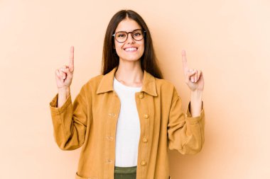 Beyazlı genç bir kadın bej arka planda izole edilmiş. İki parmağı da boş bir alan gösteriyor..