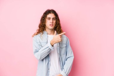 Uzun saçlı adam yalnız poz veriyor. Gülümsüyor ve işaret ediyor. Boş alanda bir şey gösteriyor..