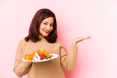 Orta yaşlı, elinde waffle tutan Latin bir kadın avuçta fotokopi alanı gösteriyor ve belinde başka bir el tutuyor..