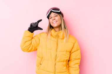 Pembe arka planda kayak kıyafetleri giyen genç beyaz bir kadın işaret parmağıyla küçük bir şey tutuyor, gülümsüyor ve kendinden emin..