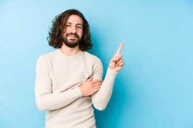 Mavi bir arka planda izole edilmiş uzun saçlı genç adam işaret parmağıyla işaret ederek gülümsüyor..