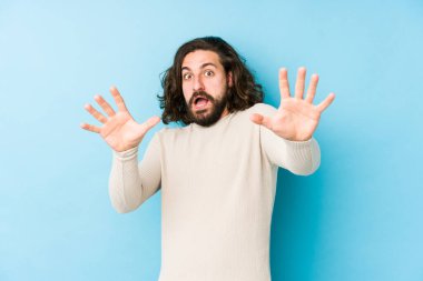 Genç, uzun saçlı bir adam, mavi arka planda izole edilmiş. Yakın bir tehlike nedeniyle şok oluyor.