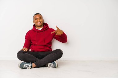 Young latin man sitting on the floor isolated person pointing by hand to a shirt copy space, proud and confident