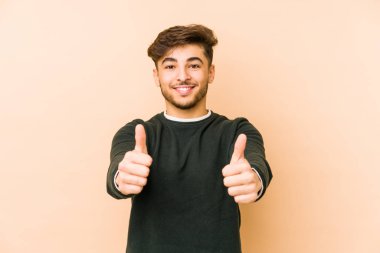 Genç Arap bir adam bej bir arka planda izole edilmiş, başparmakları havada, bir şey için tezahürat yapıyor, destek ve saygı kavramı var..