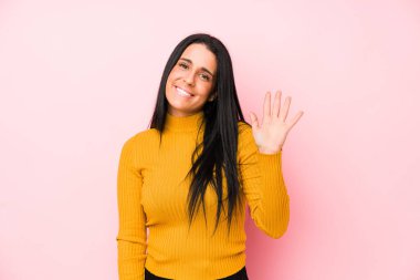 Pembe arka planda izole edilmiş genç beyaz kadın gülümseyerek beş numarayı parmaklarıyla gösteriyor..