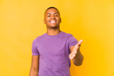 Young latin man isolated on yellow background stretching hand at camera in greeting gesture.