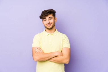 Mor bir arka planda izole edilmiş, kendinden emin, kararlılıkla kollarını kavuşturan genç bir Arap..