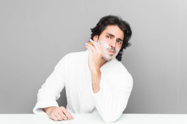 Gri bir arka planda sakalını kesen genç beyaz bir adam.