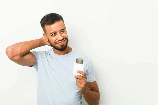 Young south-asian man holding an after shave cream touching back of head, thinking and making a choice.
