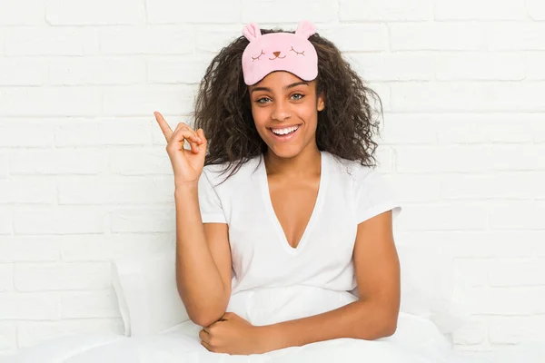 年轻的非洲裔美国女人坐在床上 戴着睡眠面具 高高兴兴地微笑着 无时无刻不带着食指 — 图库照片