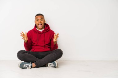 Young latin man sitting on the floor isolated celebrating a victory or success, he is surprised and shocked.