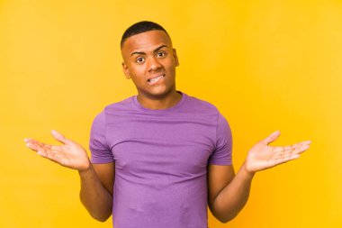 Young latin man isolated on yellow background confused and doubtful shrugging shoulders to hold a copy space.