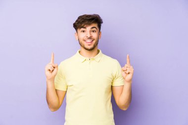Mor arka planda izole edilmiş genç bir Arap adam iki ön parmağı da boş bir alan gösteriyor..