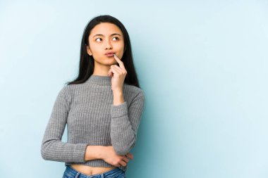 Genç Çinli bir kadın mavi bir arka planda izole edilmiş şüpheli ve şüpheci bir ifadeyle yana bakıyor..