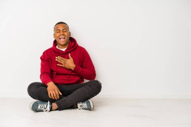 Young latin man sitting on the floor isolated laughs out loudly keeping hand on chest.