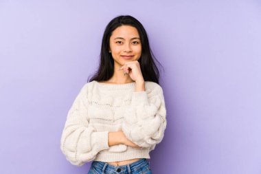 Mor arka planda izole edilmiş genç Çinli bir kadın gülümsüyor, mutlu ve kendinden emin, elleriyle çenesine dokunuyor..