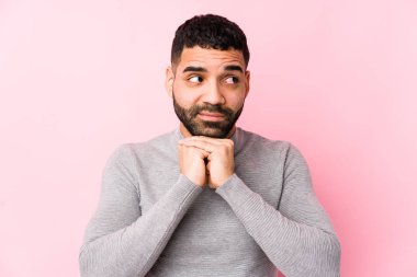 Pembe arka planı olan genç bir latin adam ellerini çenesinin altında tutuyor ve mutlu bir şekilde yan tarafa bakıyor..