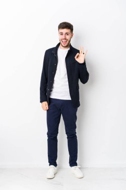 Tüm vücudu beyaz tenli genç bir adam bir göz kırpıyor ve eliyle iyi bir jest yapıyor..