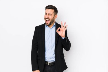 Young caucasian business man against a white background isolated winks an eye and holds an okay gesture with hand.