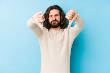 Mavi arka planda izole edilmiş genç, uzun saçlı adam baş parmağını aşağı gösteriyor ve hoşlanmadığını ifade ediyor..
