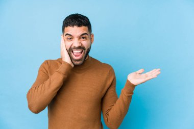 Mavi arka planı olan genç bir latin avucunun içinde fotokopi alanı tutuyor, el üstünde tutuyor. Şaşırmış ve mutlu..