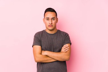 Pembe arka planda poz veren, hoşnutsuzca kaşlarını çatan, kollarını kavuşturan Latin bir adam..