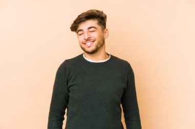 Bej bir arka planda izole edilmiş genç bir Arap, gülüyor ve gözlerini kapatıyor, rahatlamış ve mutlu hissediyor..