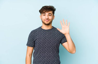 Genç Arap bir adam mavi bir arka planda izole edilmiş. Gülümsüyor beş numarayı parmaklarıyla gösteriyor..
