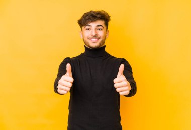 Sarı arka planda izole edilmiş genç bir Arap adam. Başparmakları havada, bir şey için tezahürat yapıyor. Destek ve saygı kavramı..