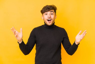 Sarı arka planda izole edilmiş genç bir Arap hoş bir sürpriz bekliyor, heyecanlı ve el kaldırıyor..