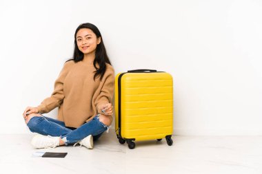 Genç Çinli gezgin bir bavulla yerde oturuyor mutlu, güler yüzlü ve neşeli..