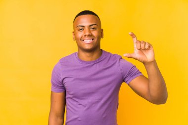 Young latin man isolated on yellow background holding something little with forefingers, smiling and confident.