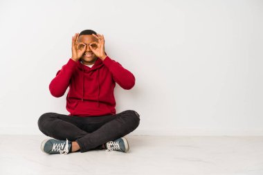 Young latin man sitting on the floor isolated showing okay sign over eyes