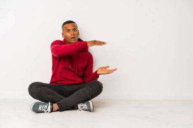 Yerde oturan genç latin adam şok olmuş ve hayrete düşmüş bir şekilde ellerinin arasında bir kopya alanı tutuyor..