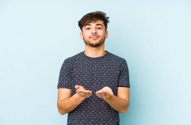 Genç Arap bir adam mavi bir arka planda izole edilmiş. Elinde avuç içi olan bir şey var ve kameraya sunuyor..