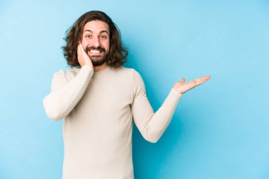 Mavi arka planda izole edilmiş genç, uzun saçlı adam avuç içinde fotokopi alanı tutuyor, el üstünde tutuyor. Şaşırmış ve mutlu..