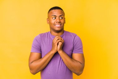 Sarı arka planda izole edilmiş genç latin adam ellerini çenesinin altında tutuyor ve mutlu bir şekilde yan tarafa bakıyor..