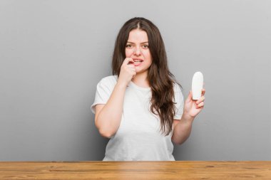 Genç ve kıvrımlı bir kadın elinde nemlendirici tutuyor tırnaklarını yiyor, gergin ve endişeli..