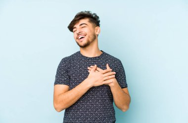 Genç Arap bir adam mavi bir arka planda izole edilmiş. Gülüyor, elini yüreğine koyuyor, mutluluk kavramını koruyor..