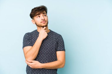 Mavi arka planda izole edilmiş genç bir Arap şüpheli ve şüpheci bir ifadeyle yana bakıyor..