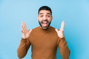 Mavi arka planda genç bir latin adam zaferi ya da başarıyı kutluyor, şaşırıyor ve şok oluyor..