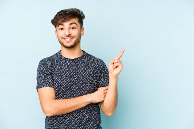 Mavi bir arka planda izole edilmiş genç Arap adam işaret parmağıyla işaret ederek gülümsüyor..