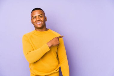 Mor arka planda izole edilmiş genç Latin adam gülümsüyor ve boş alanda bir şeyler gösteriyor..