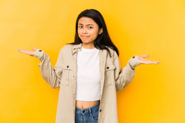 Genç Çinli bir kadın sarı arka planda izole edilmiş sorgulama sırasında omuz silkiyor ve şüphe duyuyor..