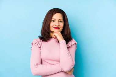 Orta yaşlı, yalnız, mutlu ve kendinden emin bir şekilde gülümseyen, elleriyle çeneye dokunan Latin kadın..