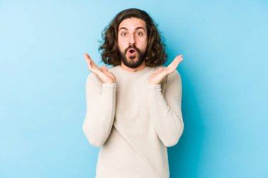 Uzun saçlı genç adam mavi bir arka planda izole edilmiş. Şaşırmış ve şok olmuş..